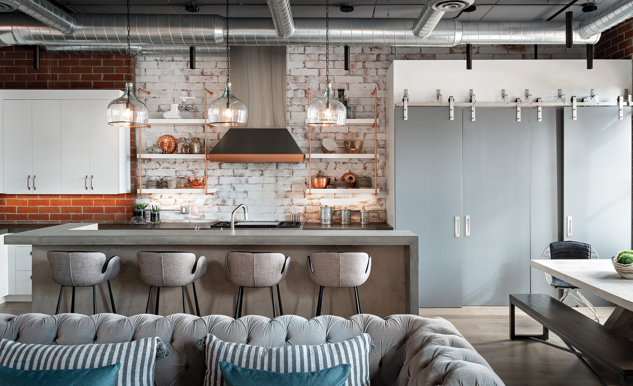 Modern open kitchen with a concrete island, four gray bar stools, exposed brick walls, glass hanging lamps, and metal ductwork ceiling.