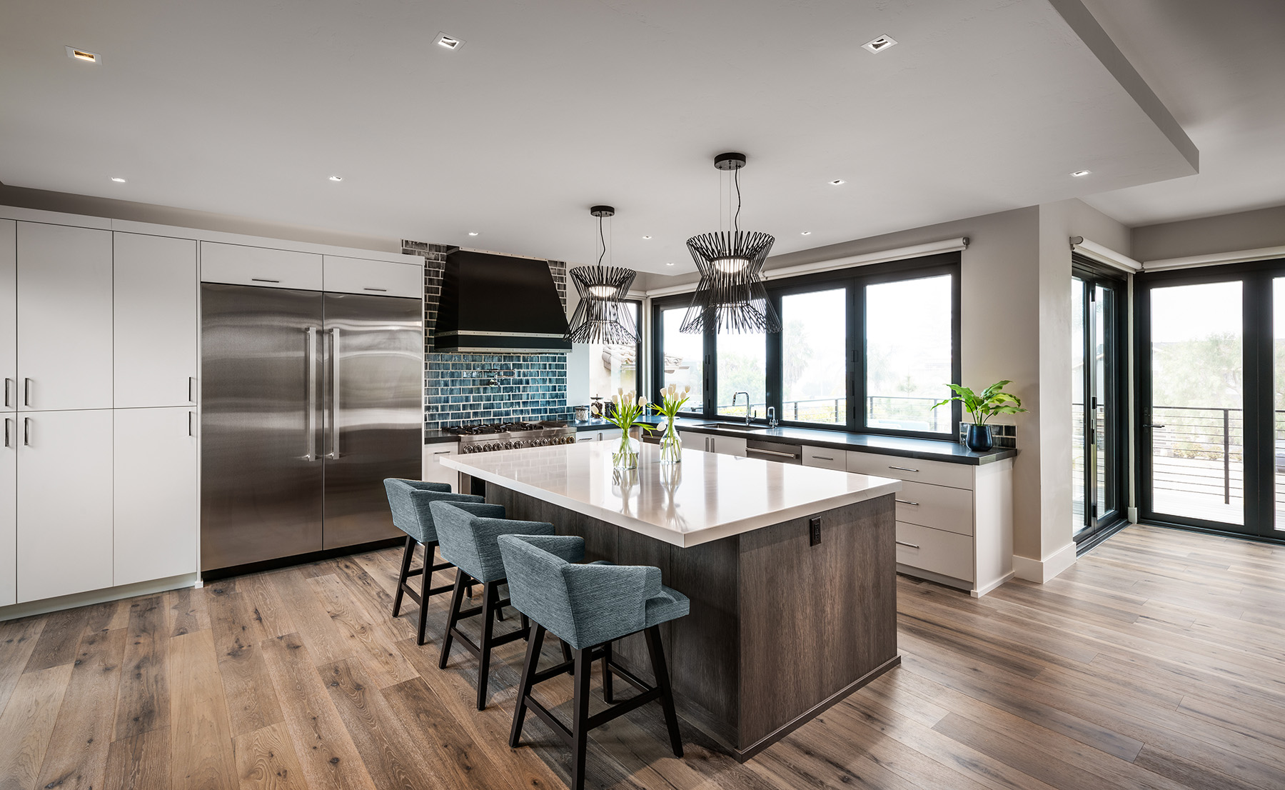 Modern kitchen with a large island featuring a white countertop, three blue upholstered bar stools, stainless steel appliances, and large windows letting in natural light.