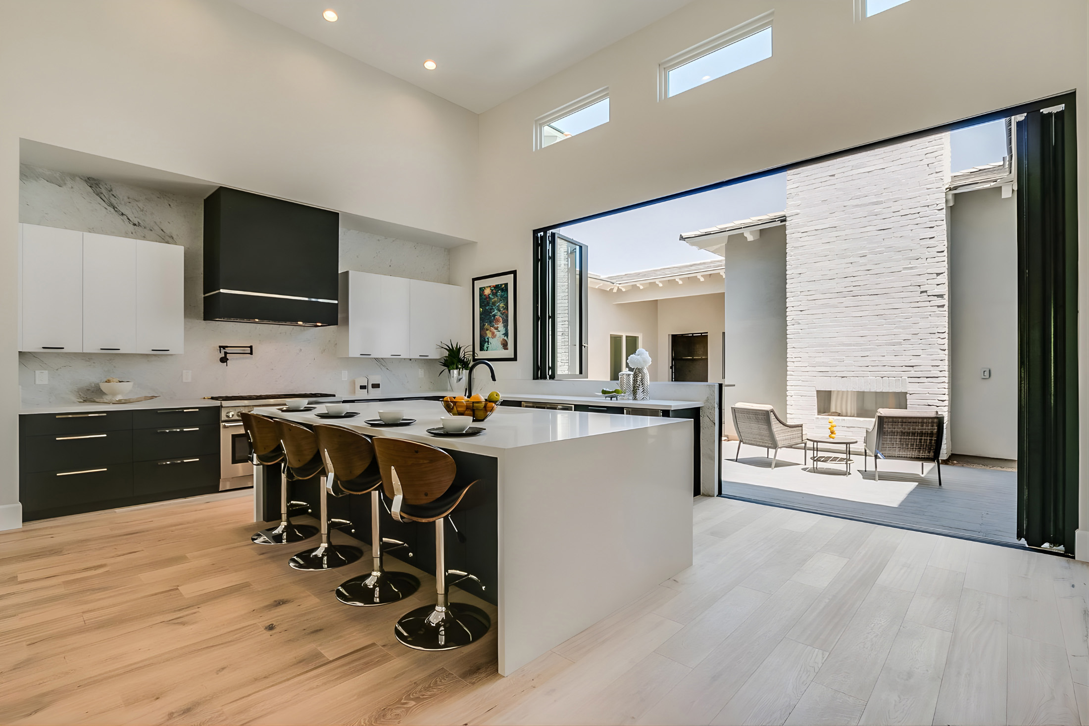 Modern kitchen with white and black cabinetry, large island with four wooden bar stools, and open sliding doors leading to outdoor seating area with fireplace.