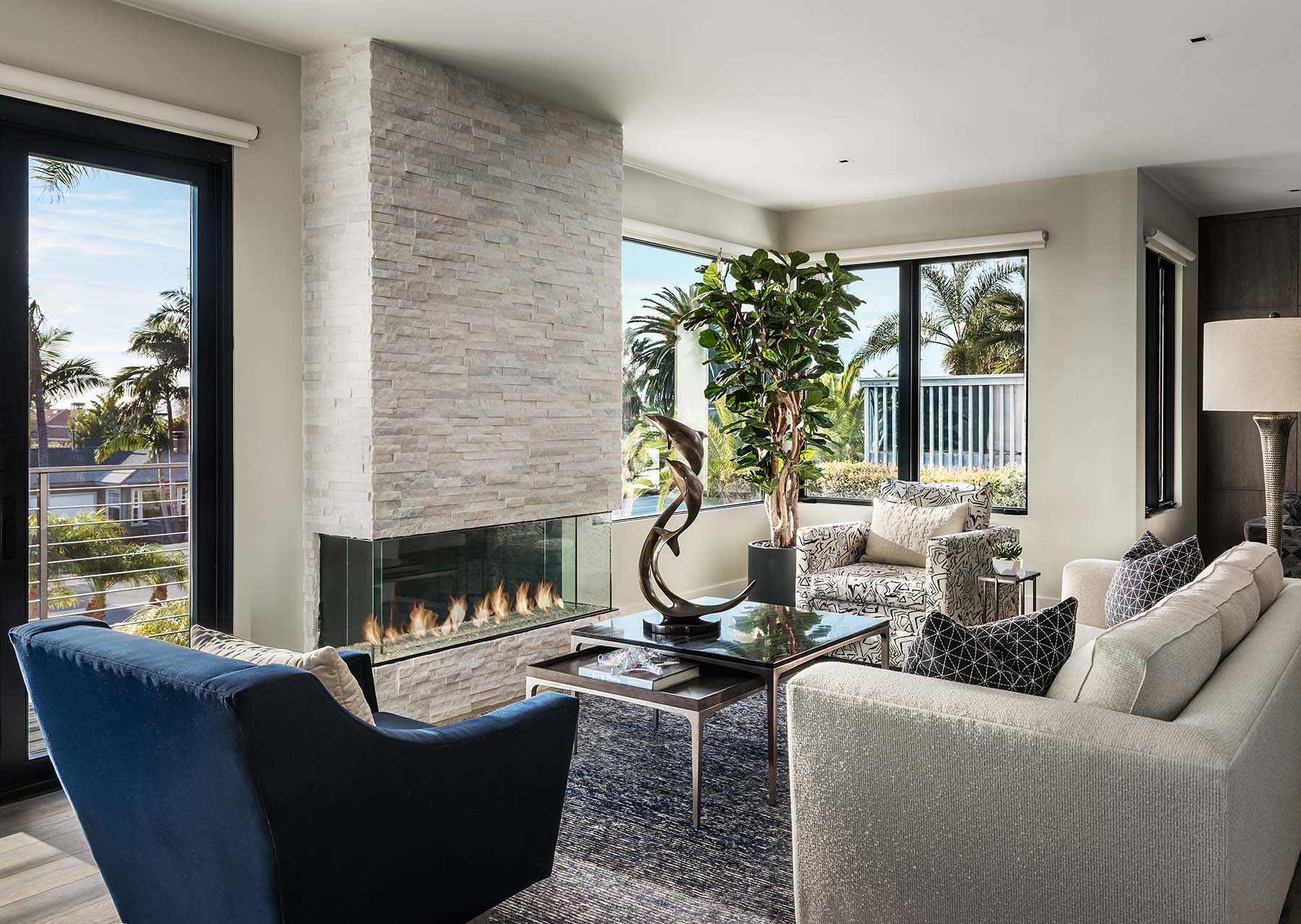 Modern living room with a stone fireplace, navy and beige seating, glass coffee table with sculpture, and large windows showing palm trees outside.