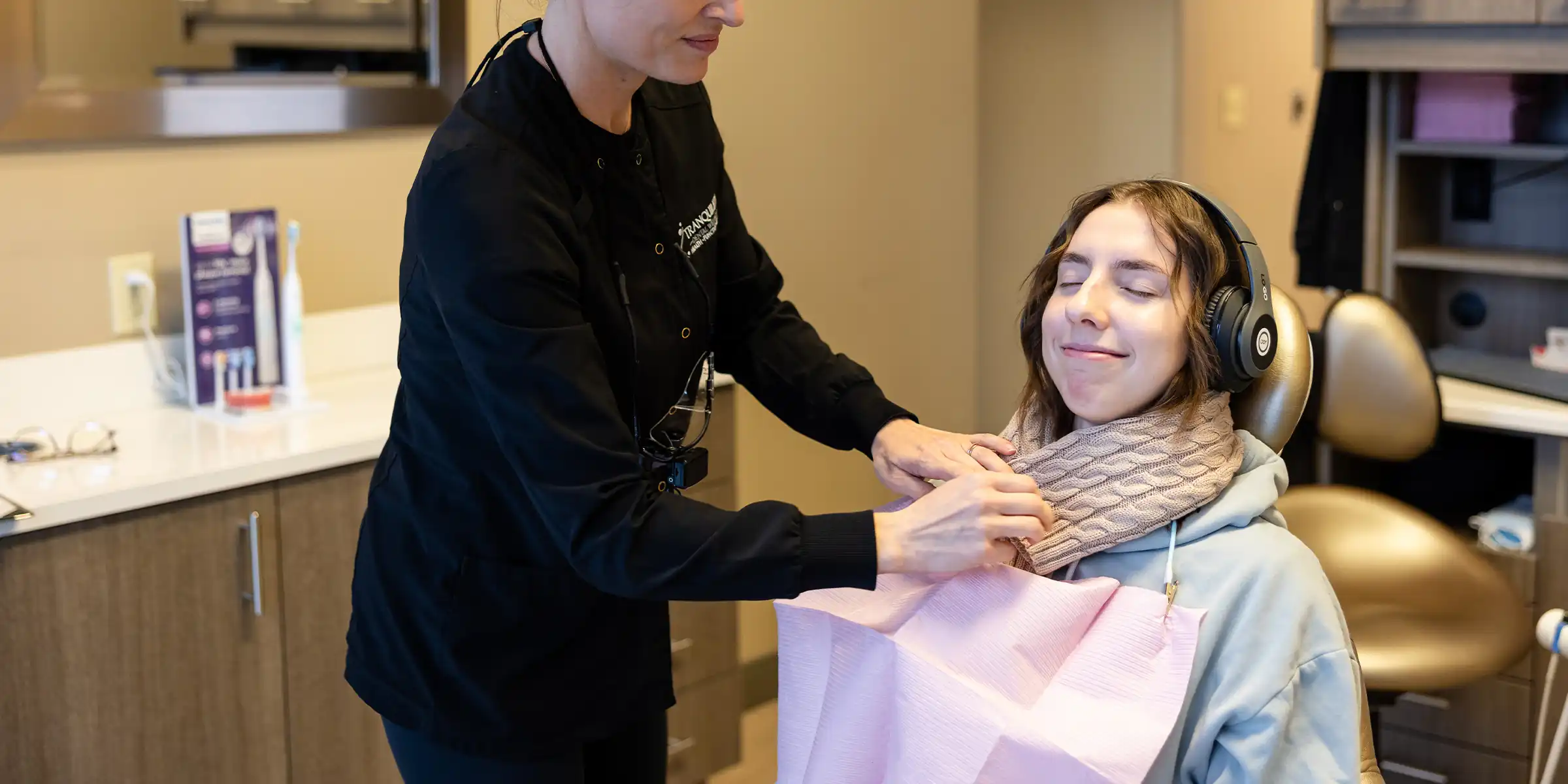 One of Tranquility Dental Wellness' hygienist placing a warm herbal neck roll around a patients neck as the patient relaxes in a dental chair wearing headphones.