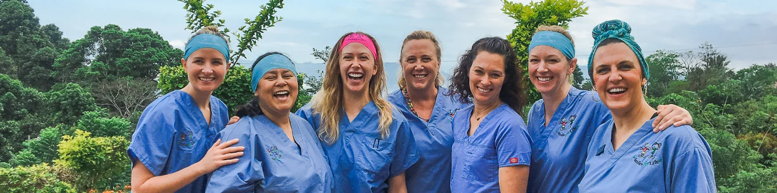 Group of seven dental professionals wearing blue scrubs and headbands smiling, standing close together outdoors with green trees in the background.