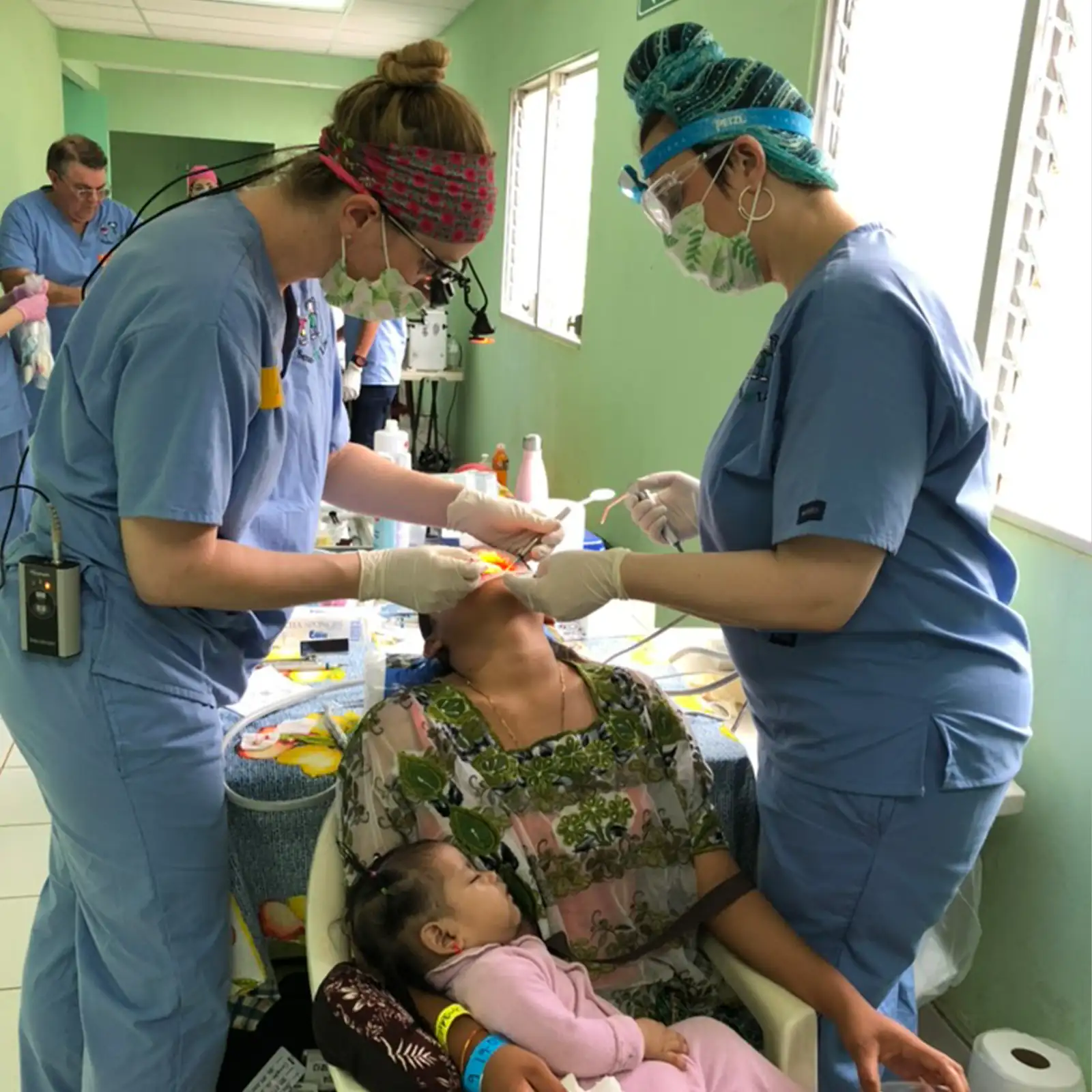Two dental professionals in blue scrubs and masks treating a patient seated in a chair holding a sleeping child.