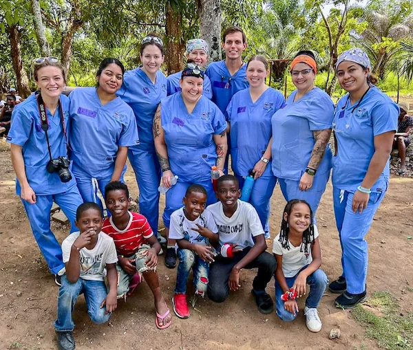 Group of Tranquility Dental workers in blue scrubs posing outdoors with smiling children on a charity mission trip.