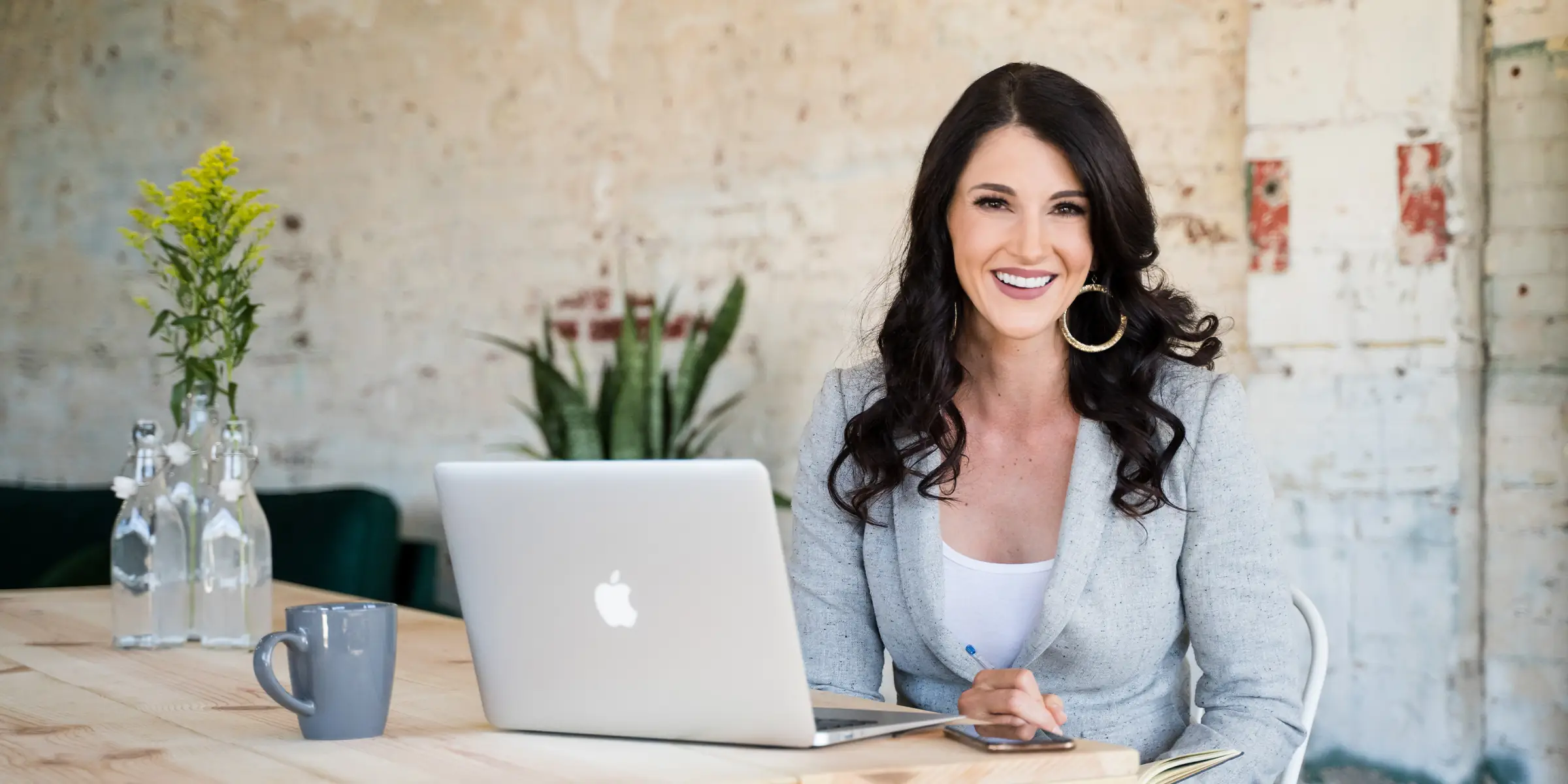 Smiling woman with dark curly hair sitting at wooden table with laptop, notebook, and coffee mug in bright casual office.