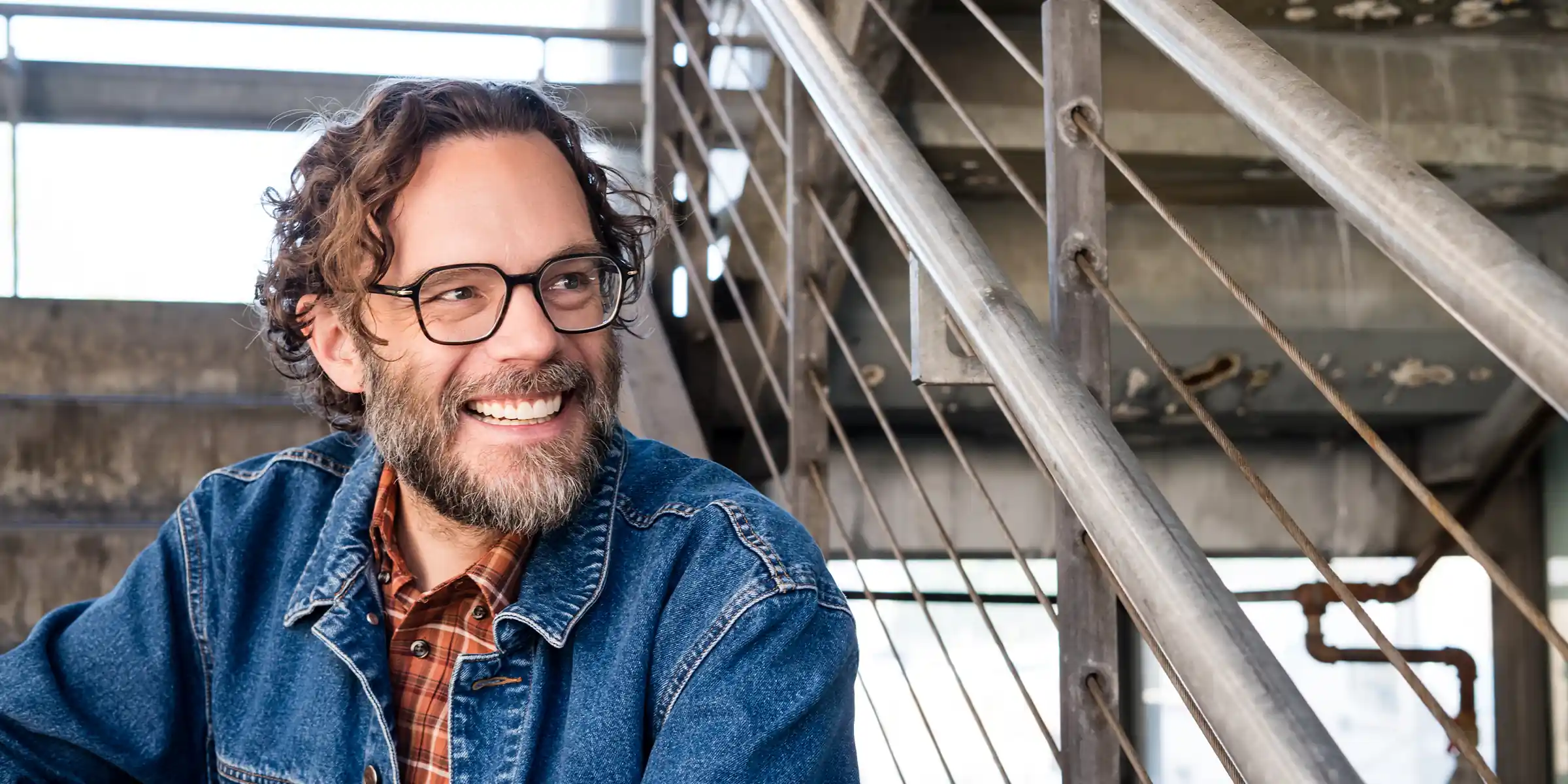 Smiling man with curly hair, beard, glasses, wearing a denim jacket and plaid shirt, sitting near metal railing indoors.