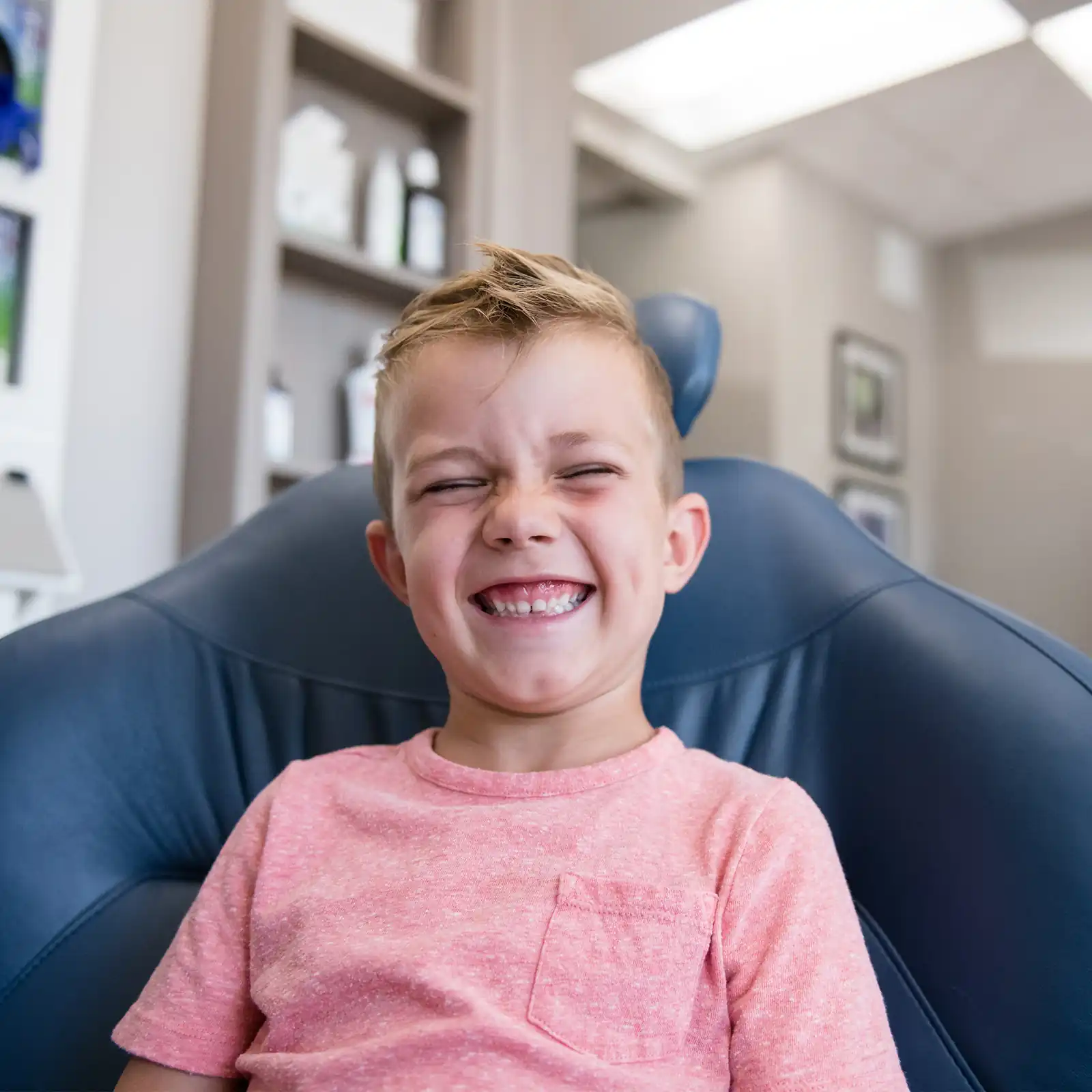 Smiling young boy with closed eyes sitting in a blue chair at the dentist's office, wearing a pink t-shirt.