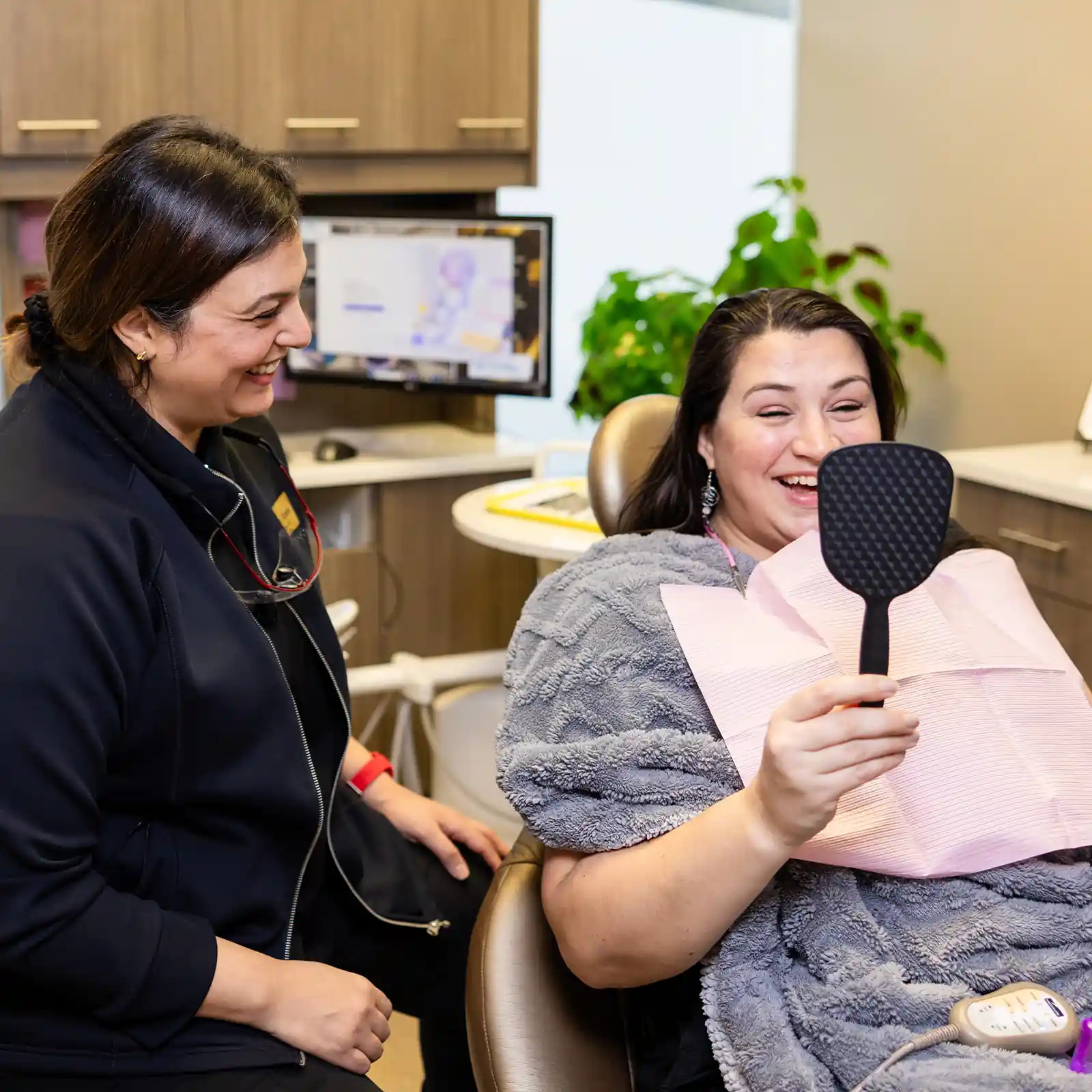 Smiling woman in dental chair holding a hand mirror while a female dental professional looks on happily.
