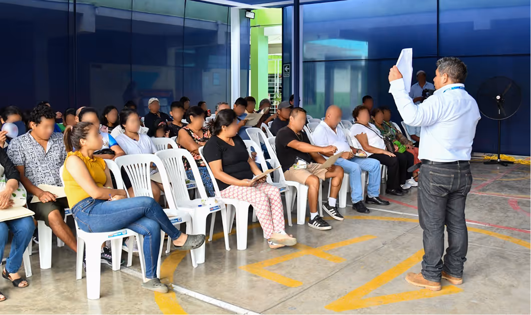 Persona de pie sosteniendo papeles mientras un grupo de personas sentadas escucha en una sala con paredes azules.