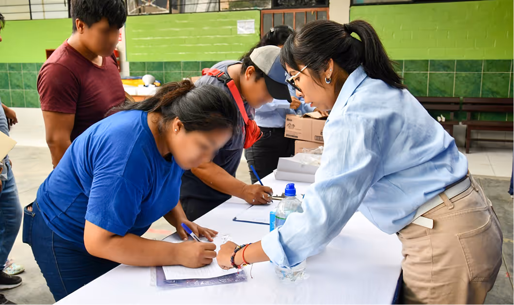 Personas firmando documentos sobre una mesa blanca en un evento con fondo verde.