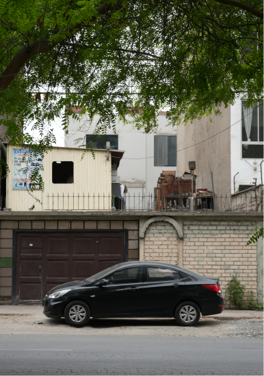 Auto sedán negro estacionado frente a una casa con muro de ladrillos y árboles verdes arriba.
