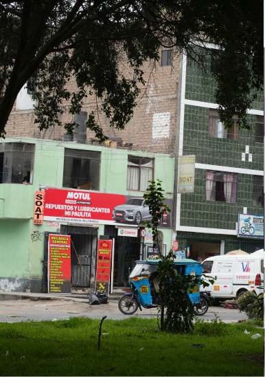Vista de edificios comerciales y residenciales con un mototaxi azul parqueado frente a una tienda de repuestos y lubricantes en una calle urbana.