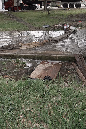 Un parche de césped embarrado y un camino de cemento húmedo y sucio cubierto con un largo trozo de tela y algunas tablas, con un gran camión y gente al fondo.