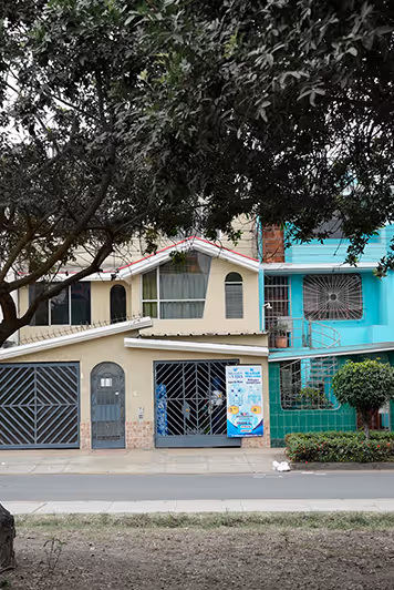 Vista de dos casas urbanas adyacentes con portones de metal, una de color beige con una pancarta que anuncia el suministro de agua, la otra de color turquesa con un pequeño jardín delantero y un árbol.