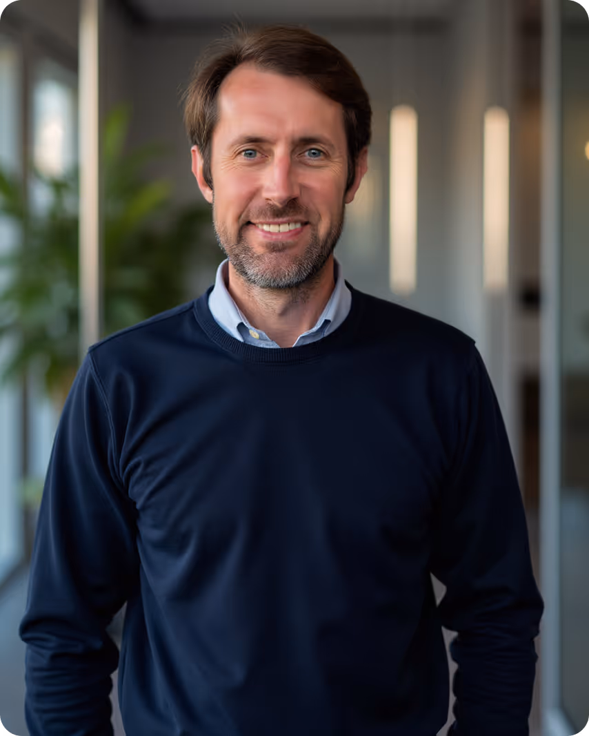 Smiling middle-aged man with short brown hair and a beard wearing a navy sweater over a light blue collared shirt in an office corridor.