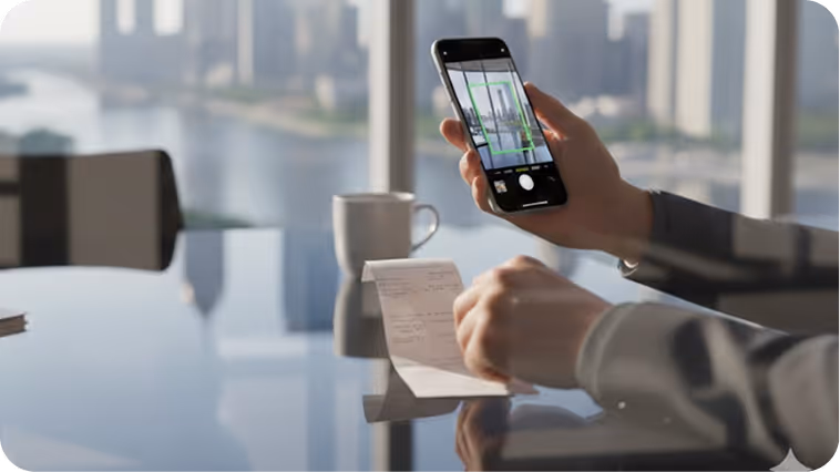 Person scanning a receipt with a smartphone over a reflective glass table in a modern office with city skyline view.
