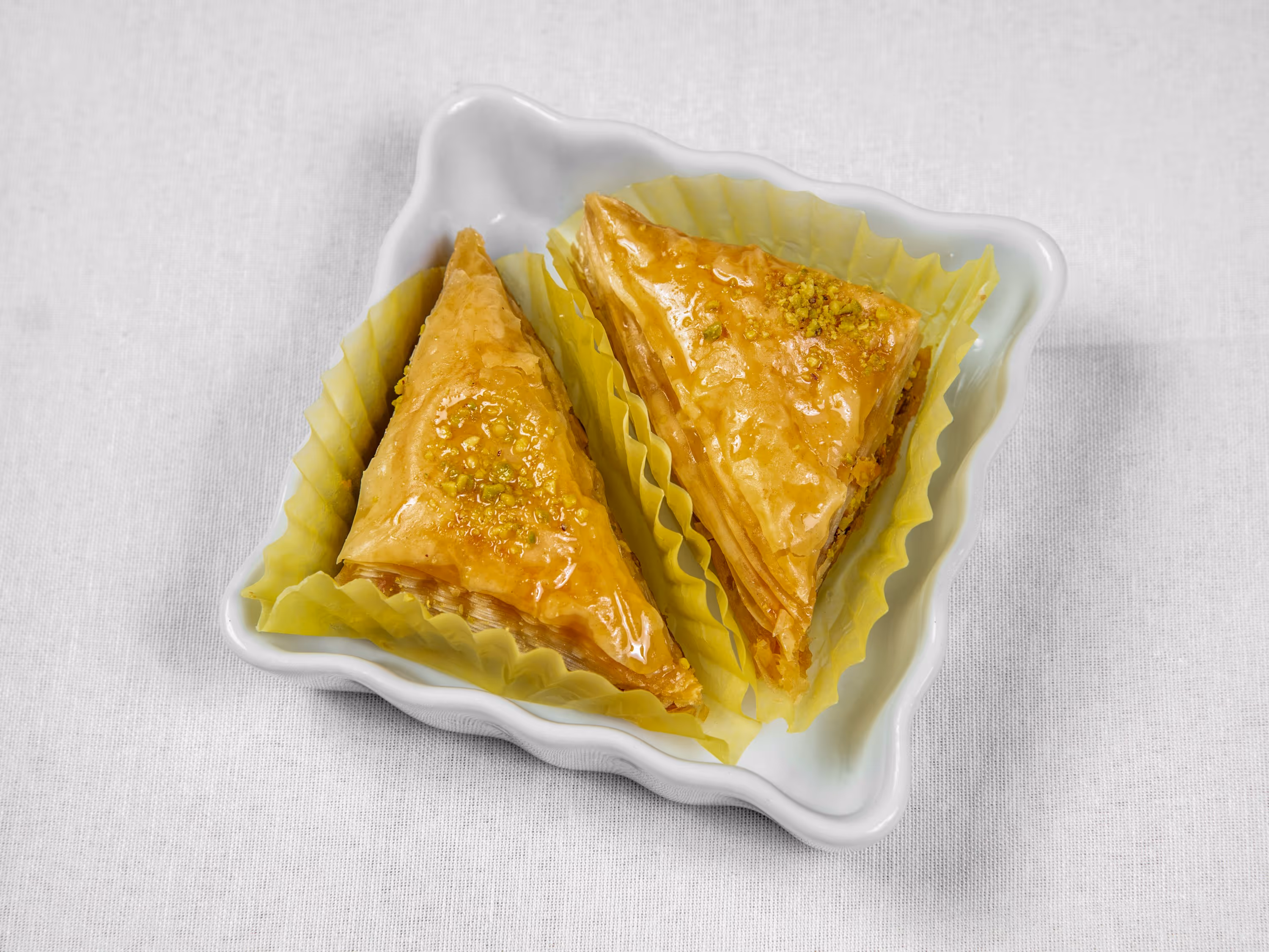Two triangular pieces of golden baklava with crushed pistachio on top, served in yellow paper cups on a white ceramic dish.