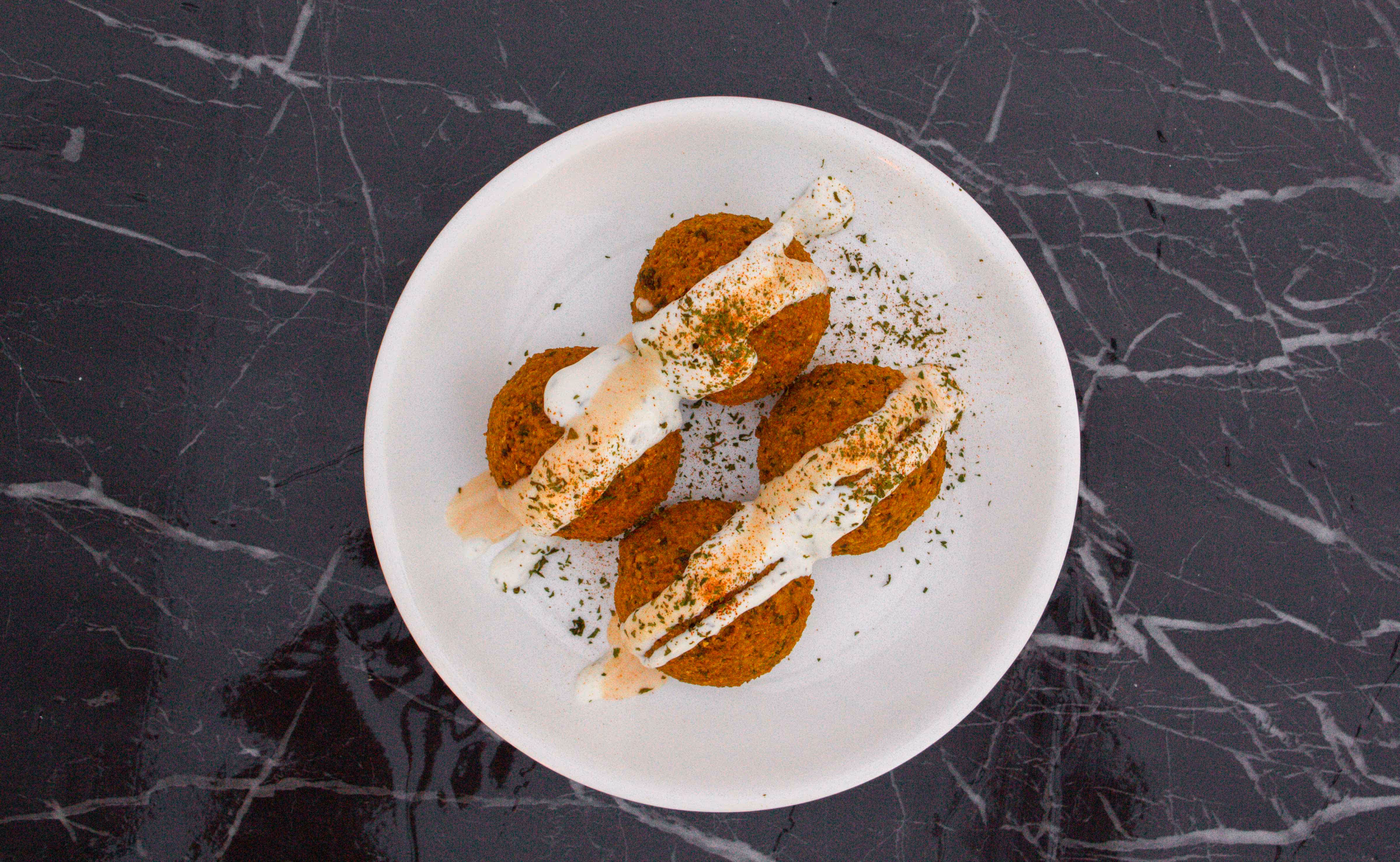 A white plate with four falafel balls topped with drizzled white sauce and sprinkled herbs on a dark marble surface.