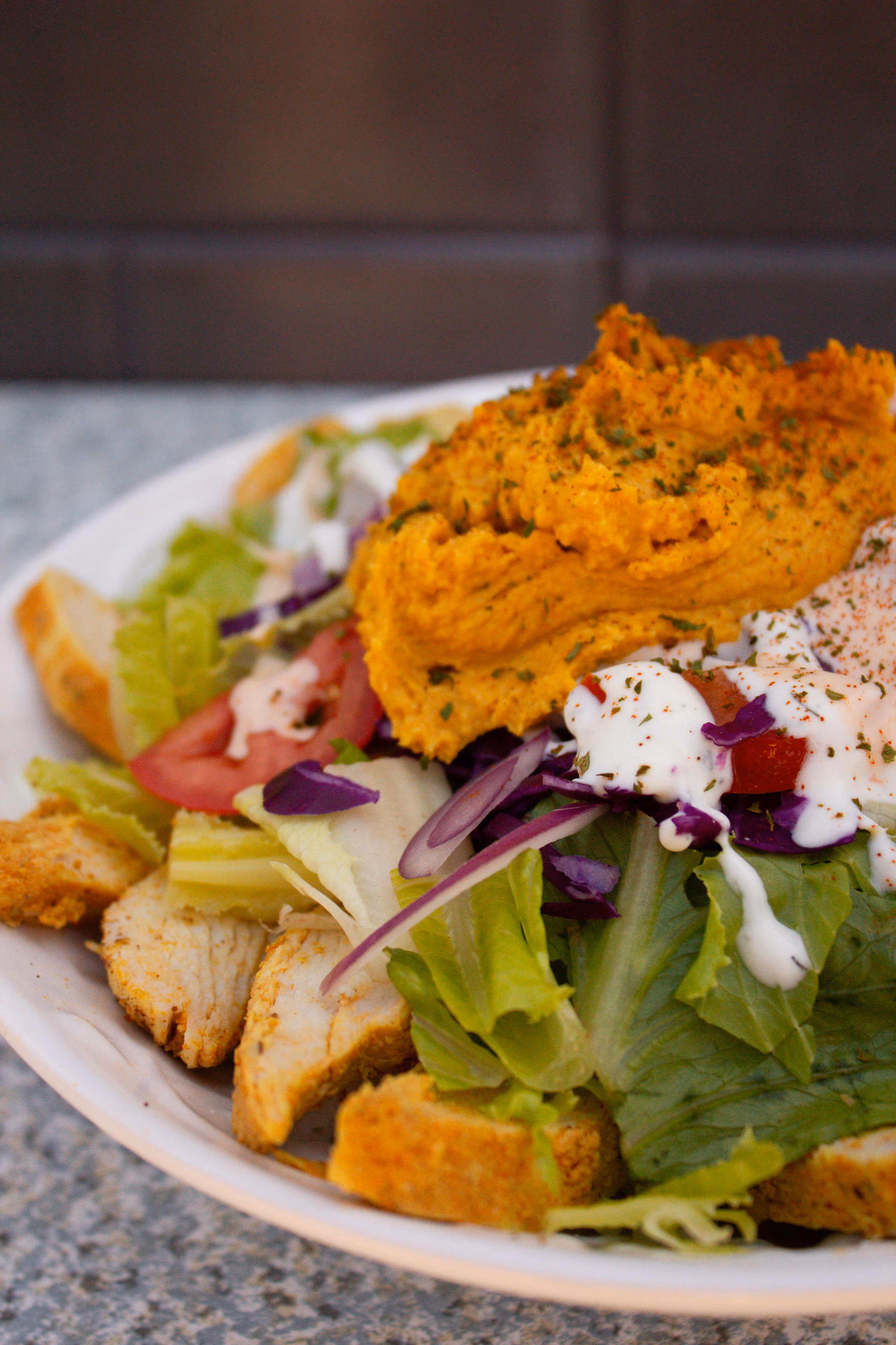 Salad with lettuce, tomato, grilled chicken, croutons, shredded purple cabbage, a dollop of orange hummus, and drizzled with creamy dressing.