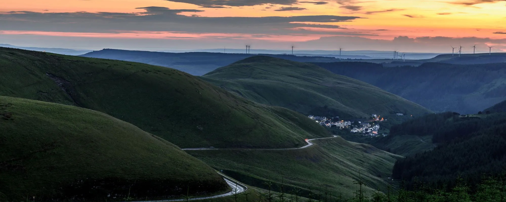 Sunset over rolling green hills with winding road and distant wind turbines