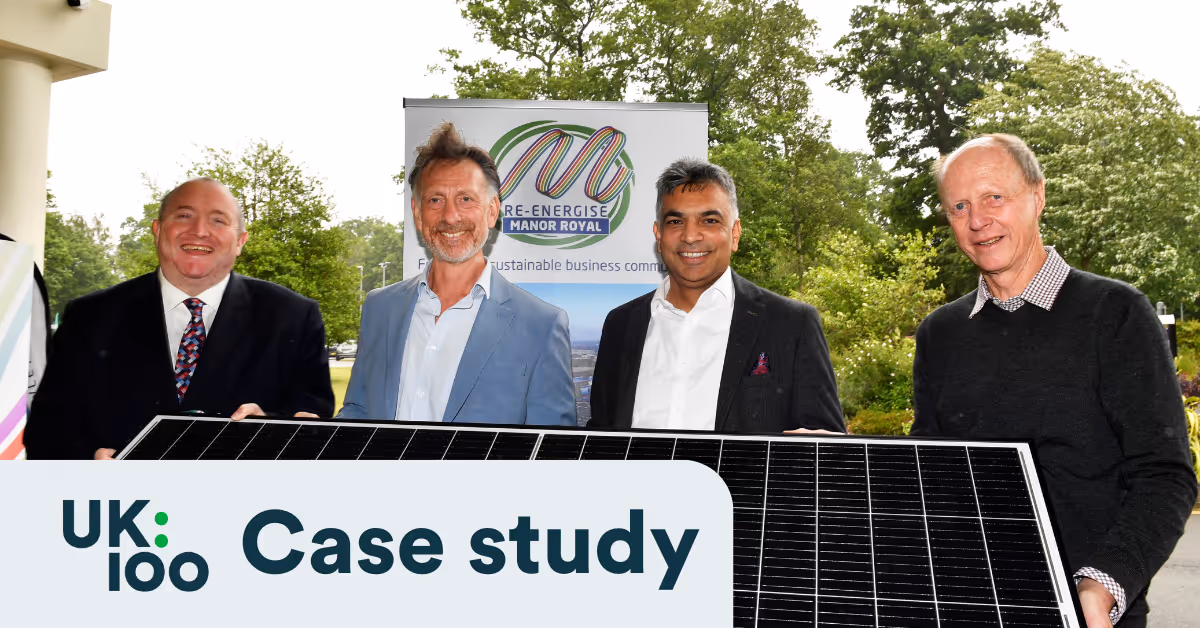 Four men standing outdoors holding a solar panel in front of a Re-Energise Manor Royal banner, with the UK100 Case Study logo overlaid in the bottom-left corner
