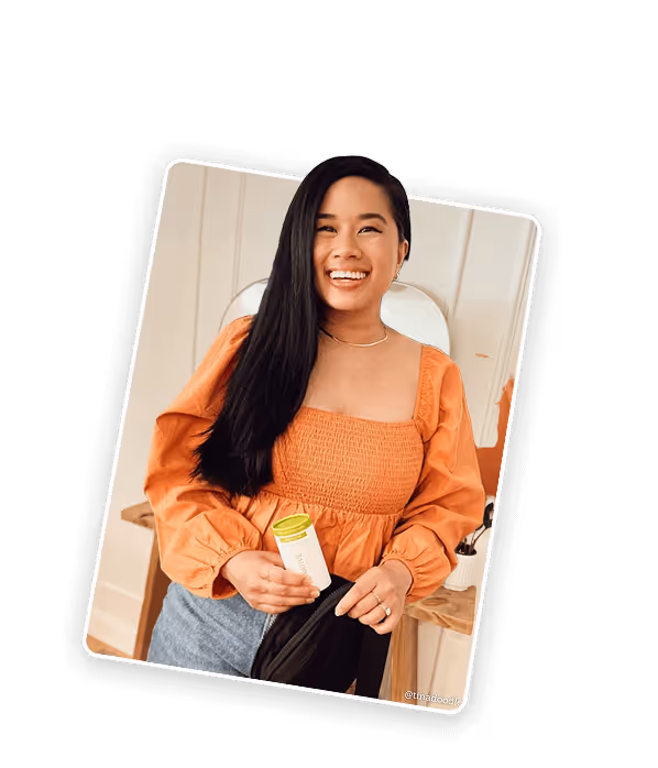 Smiling woman with long black hair wearing an orange blouse holding a small container, standing indoors near a mirror and wooden table.