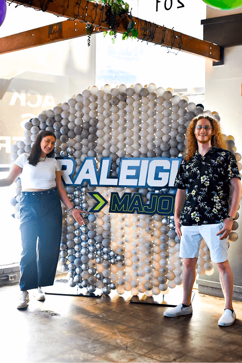 Two people smiling and posing beside a large balloon backdrop displaying the text 'RALEIGH MAJOR' in a bright indoor setting.