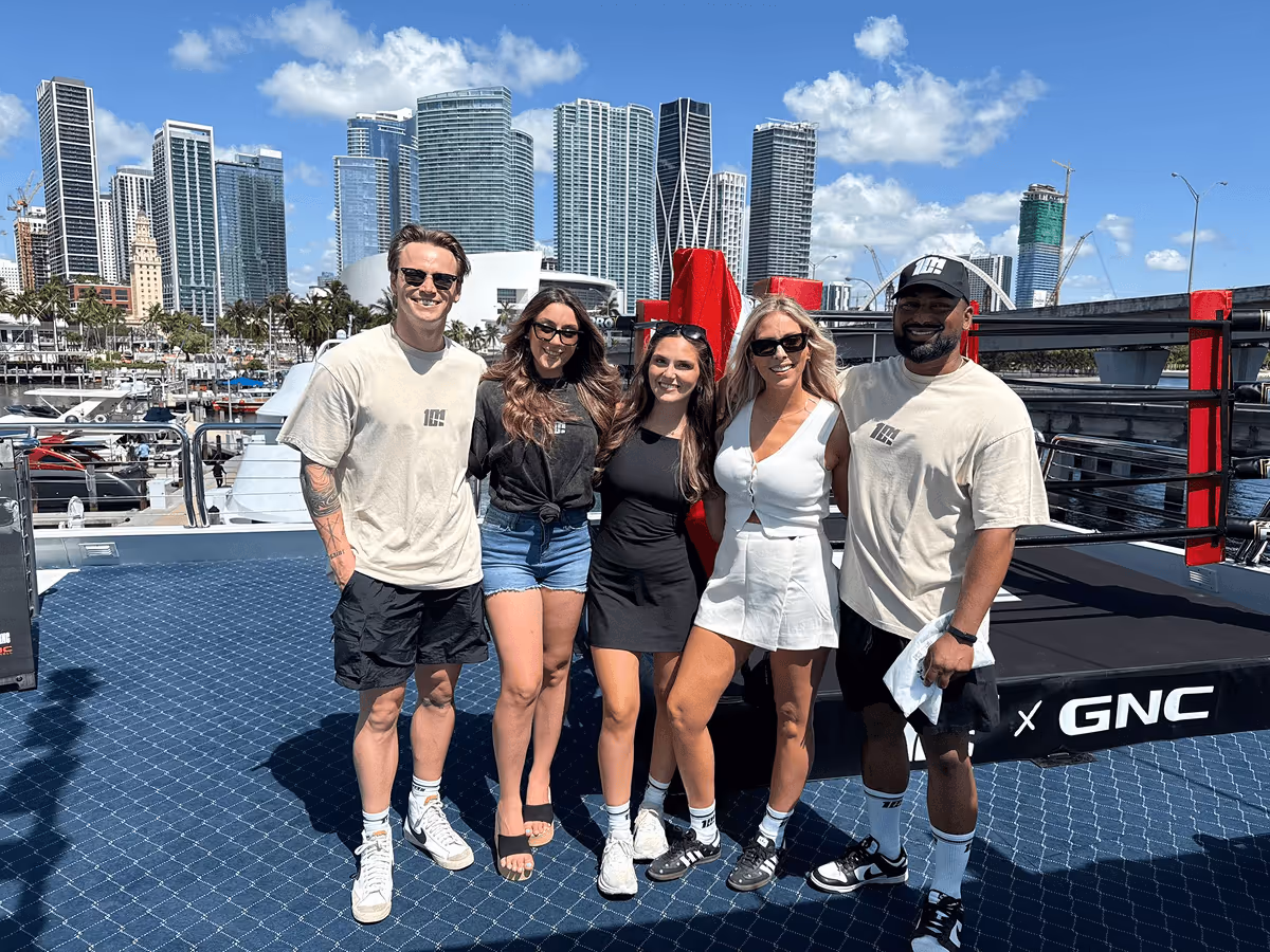 Five smiling people standing on a dock with a city skyline and yachts in the background on a sunny day.