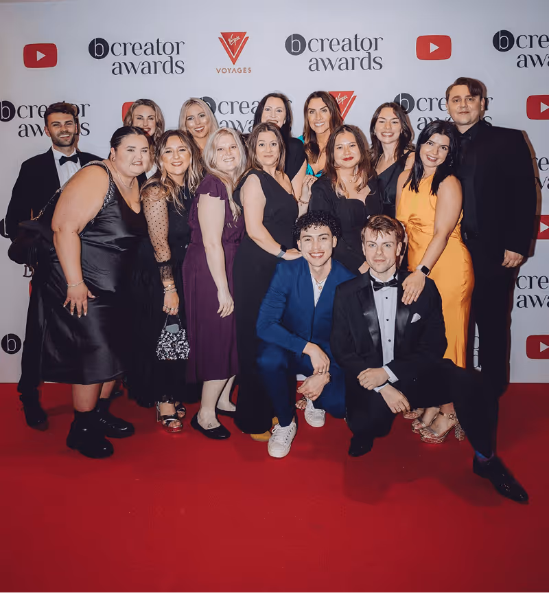 Group of fifteen people dressed in formal attire posing on a red carpet in front of a backdrop with YouTube and Creator Awards logos.