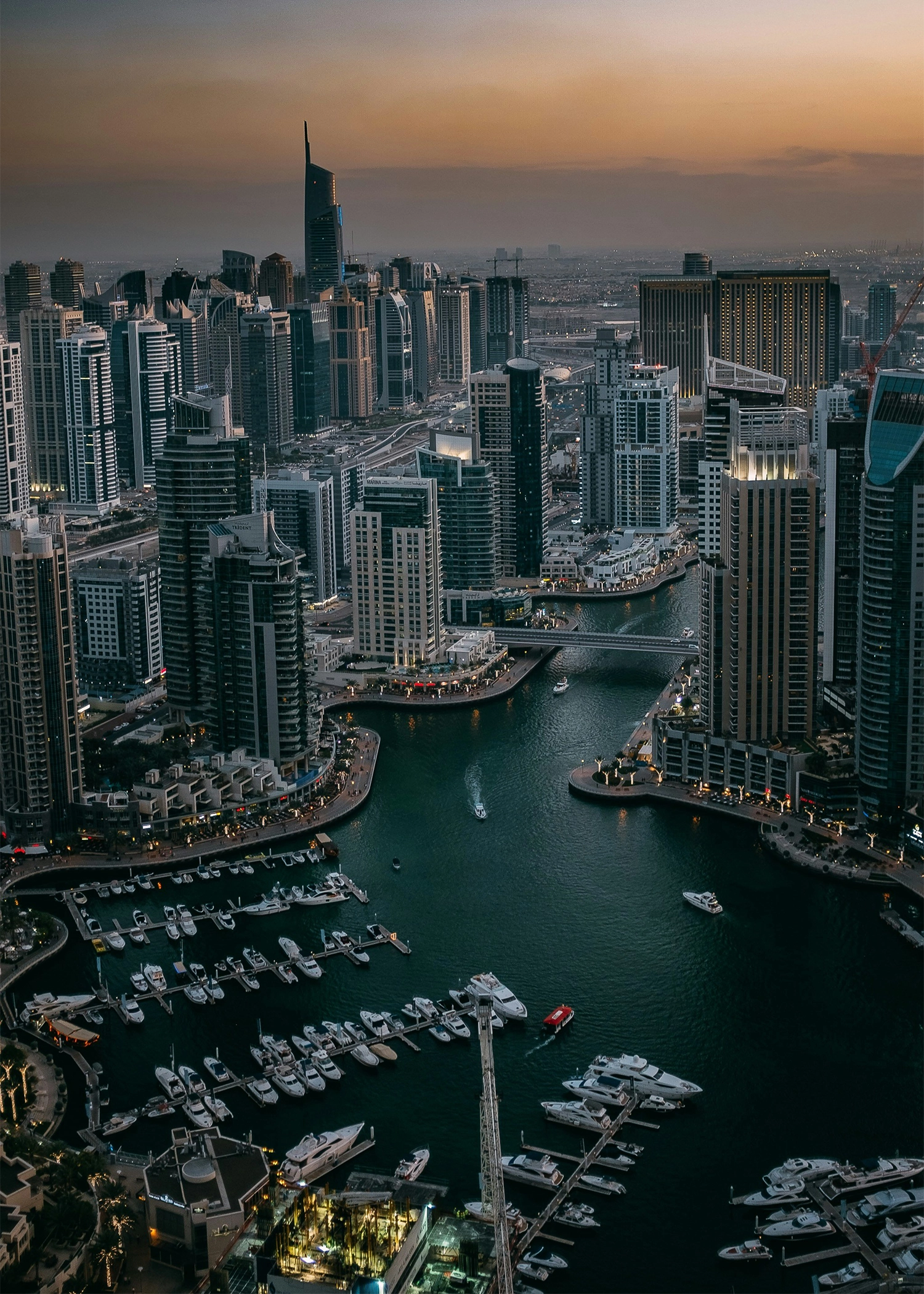 Aerial view of a marina with numerous yachts and boats docked, surrounded by tall modern skyscrapers at dusk.