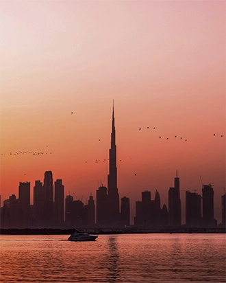 Silhouette of a city skyline with Burj Khalifa and birds flying at sunset over calm water with a boat.