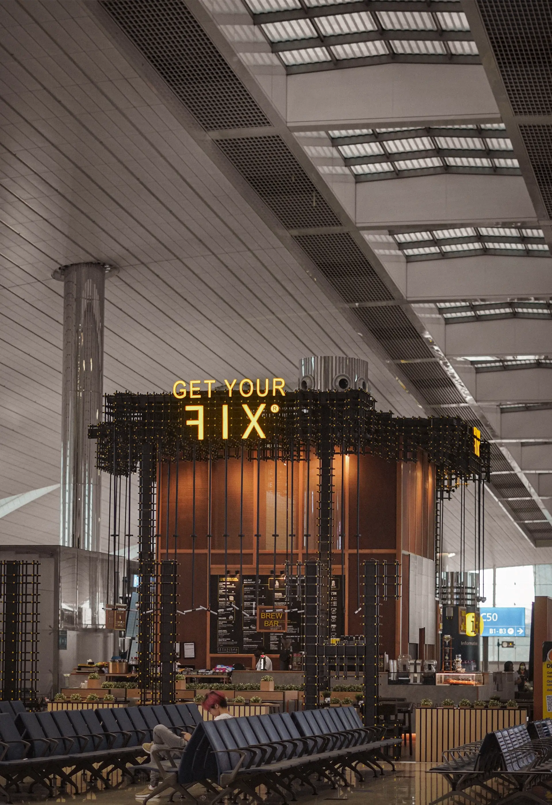 Modern coffee shop kiosk named 'Get Your Fix' inside a spacious airport terminal with rows of empty seats and a person sitting nearby.