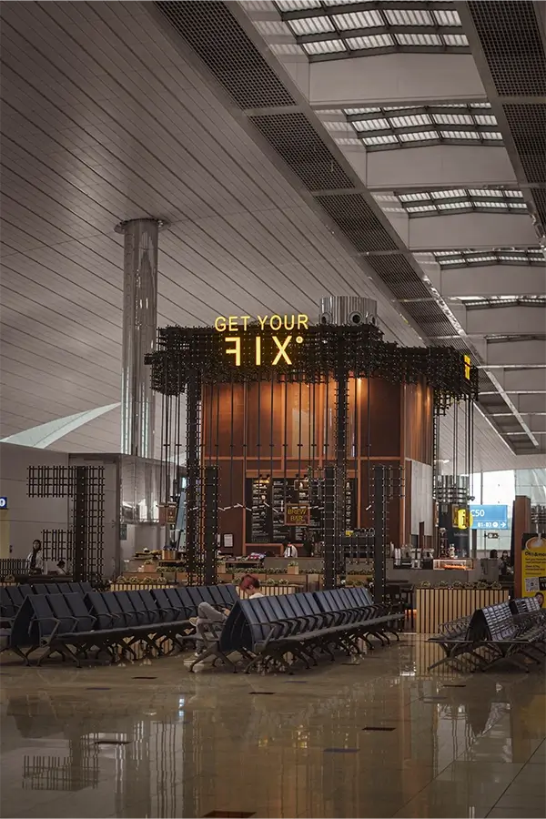 Modern airport waiting area with rows of black seats and a coffee kiosk illuminated with yellow signage reading 'GET YOUR FIX'.