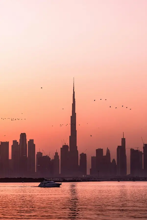 Silhouette of Dubai skyline with Burj Khalifa at sunset and a yacht on calm water.