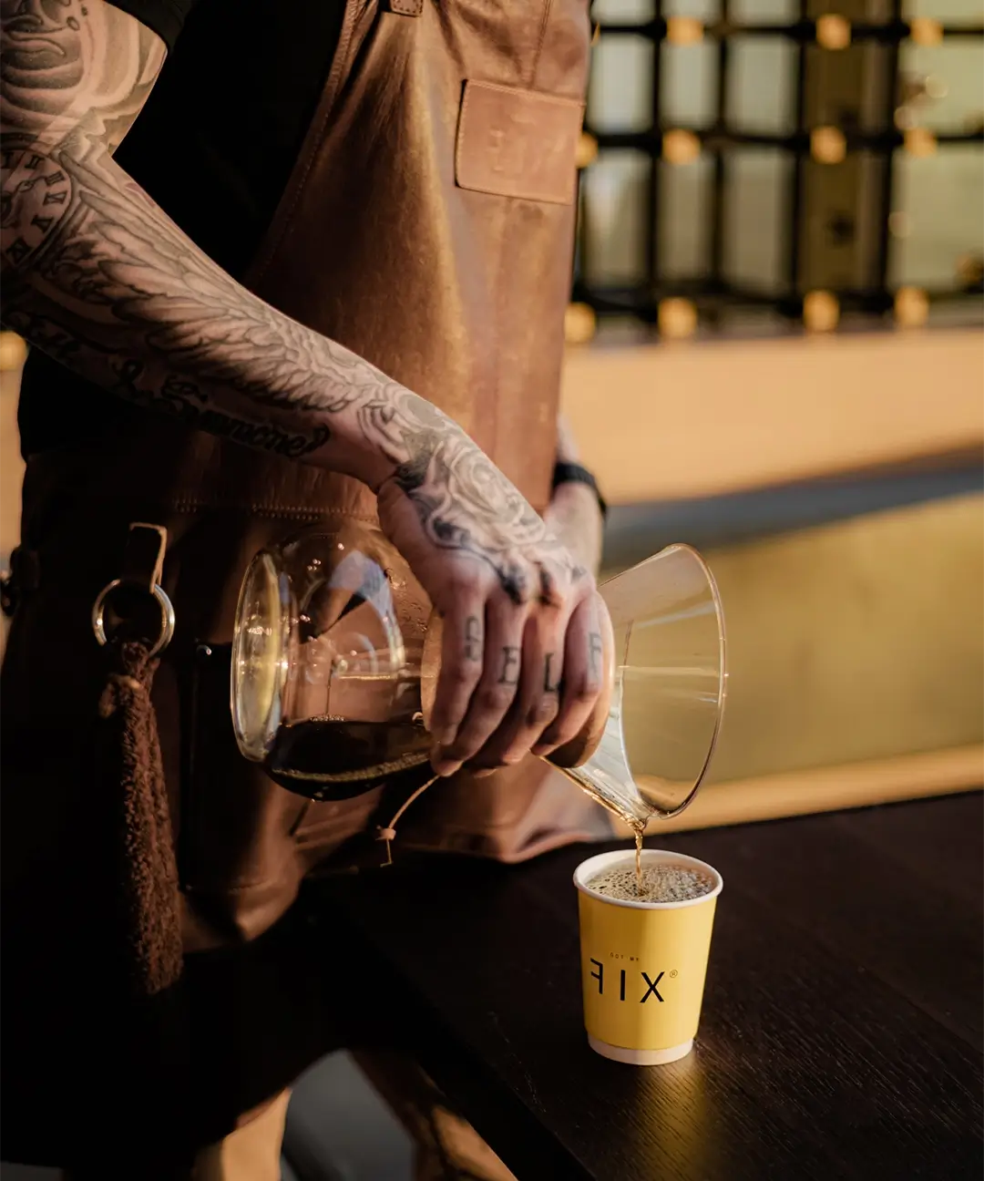 Tattooed person wearing a brown leather apron pouring coffee from a glass Chemex into a yellow paper cup on a dark wooden table.