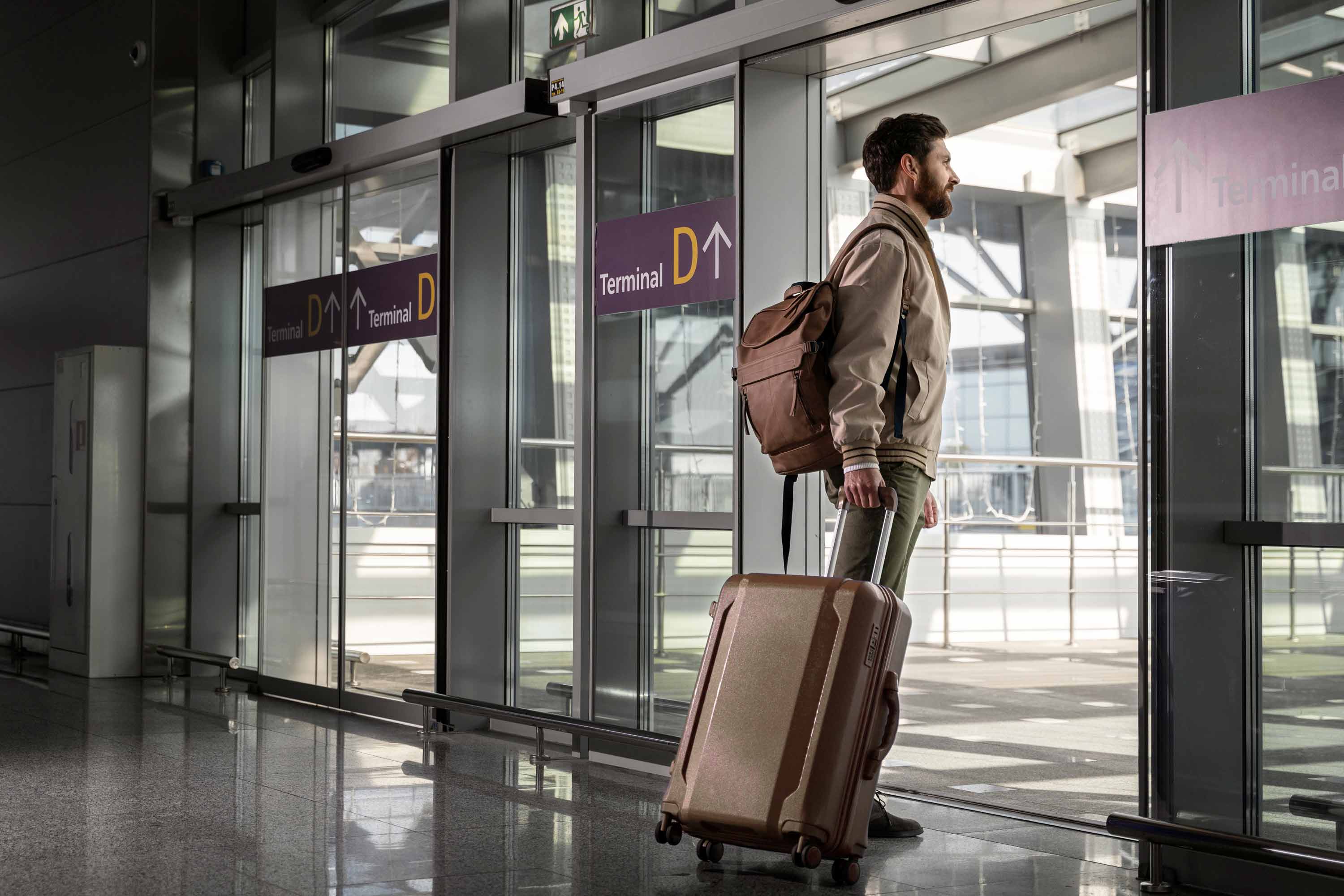 A person stands near glass doors, holding a suitcase and wearing a backpack.
The glass doors reflect the interior of the terminal, creating a sense of spaciousness.
Signages indicate “Terminal D,” with arrows pointing in that direction.
The polished, reflective floor enhances the overall brightness of the area.
