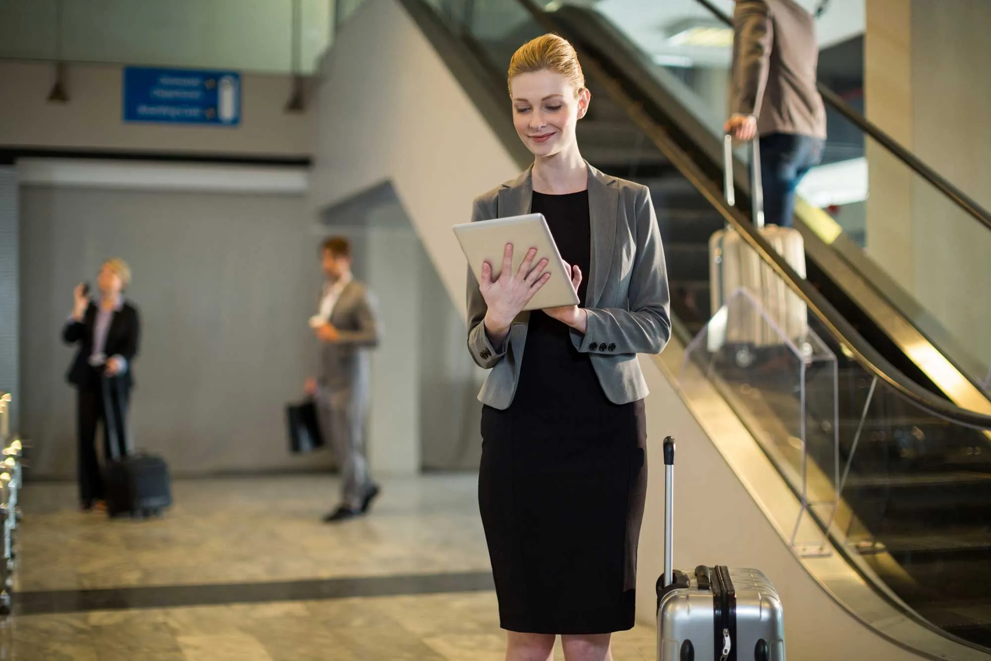 An airport setting area: with a female in formal attire holding a tablet and standing next to a suitcase.
Two other individuals in the background: one on the phone near luggage, and another ascending an escalator.
Architectural elements include an escalator, glass panels, and metallic railings.
