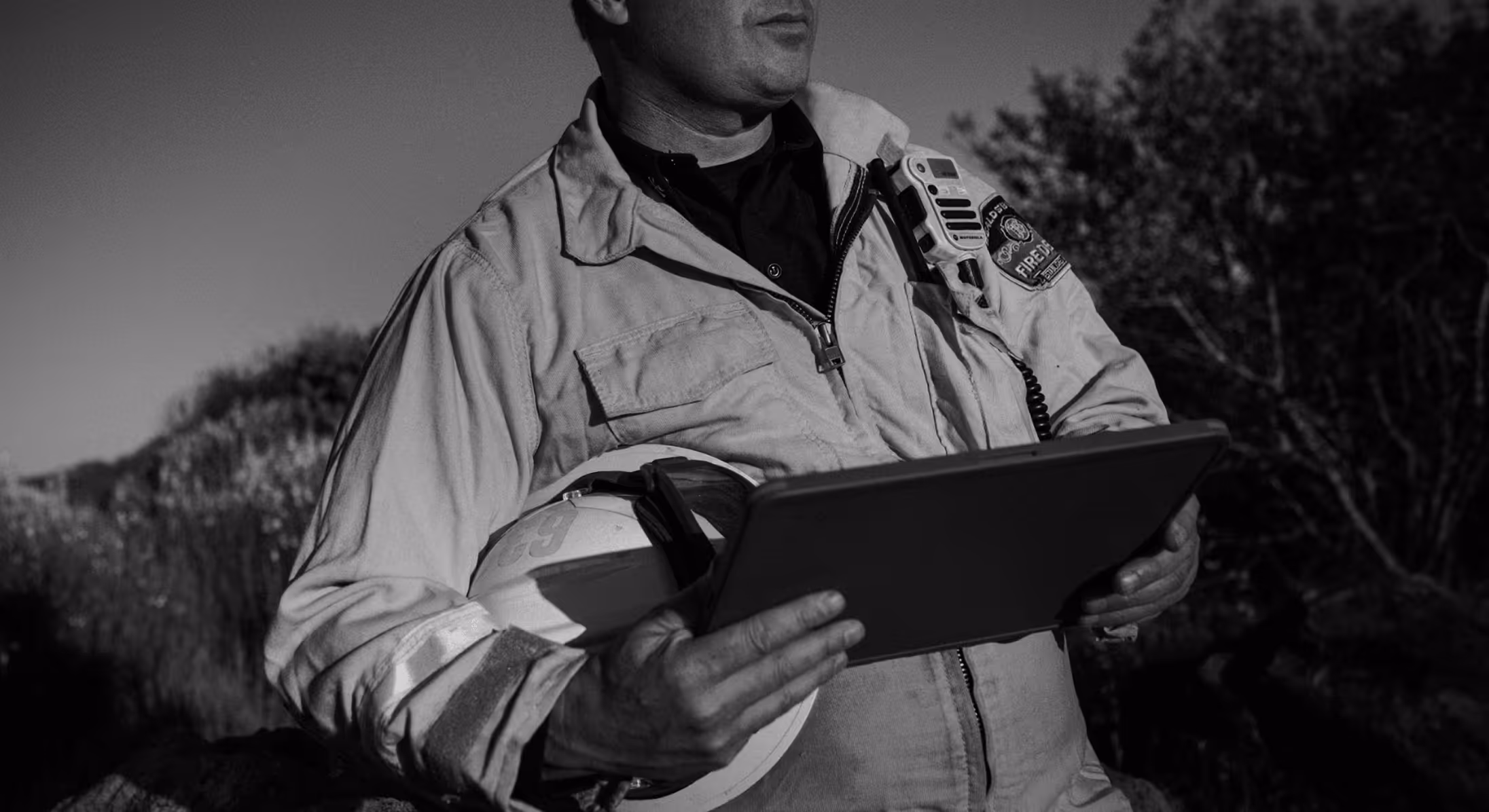 Firefighter holding a helmet and tablet outdoors wearing a radio on his jacket.