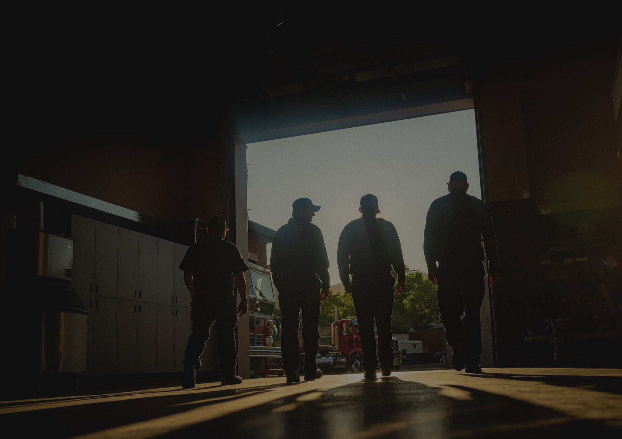 Silhouettes of four firefighters walking inside a fire station toward the open door at sunset.