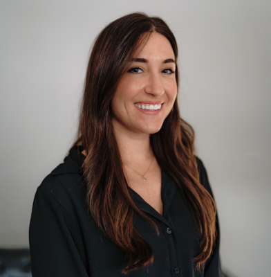 Smiling woman with light curly hair and hoop earrings wearing a black scrub top against a neutral background.