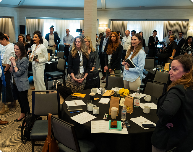Group of people standing and socializing in a conference room with round tables, notebooks, coffee cups, and water bottles.