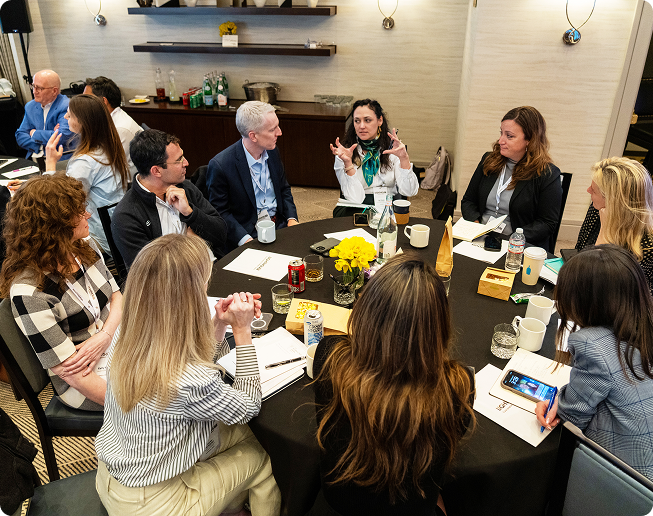 Group of people seated around a round table in a meeting room engaged in discussion, with notebooks, drinks, and yellow flowers on the table.