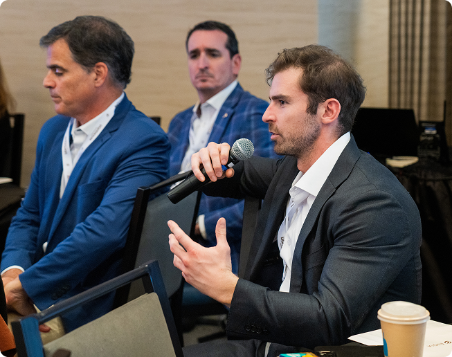 Man in a dark suit speaking into a microphone during a conference, with two men seated in the background.