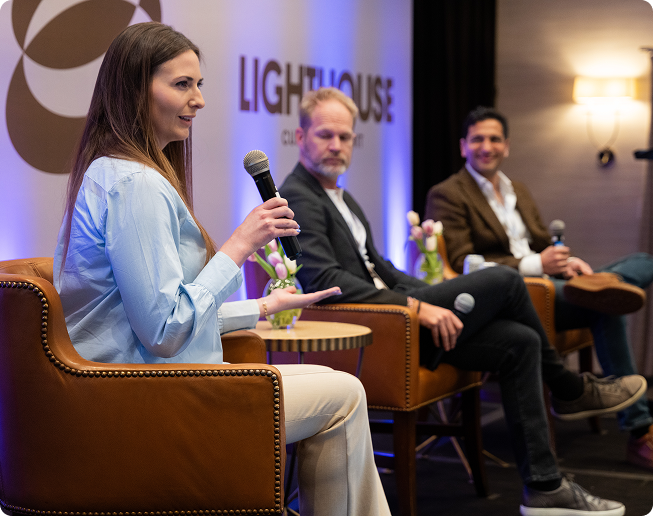 Three panelists seated on stage in brown leather chairs; a woman in light blue shirt is speaking into a microphone.