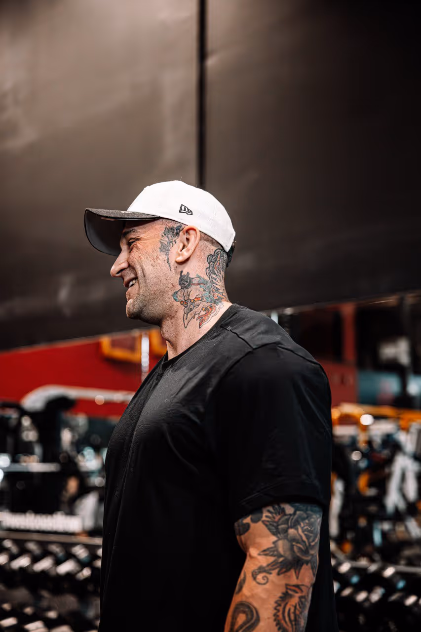 Smiling man with tattoos on neck and arm wearing a white cap and black t-shirt in a gym.