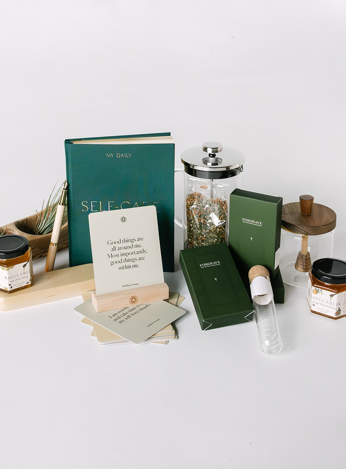 Collection of self-care items including a green journal titled 'My Daily Self-Care', affirmation cards, two jars of Anellabees honey, tea in a French press, green Forgrave boxes, and a glass honey pot with wooden dipper on a white surface.