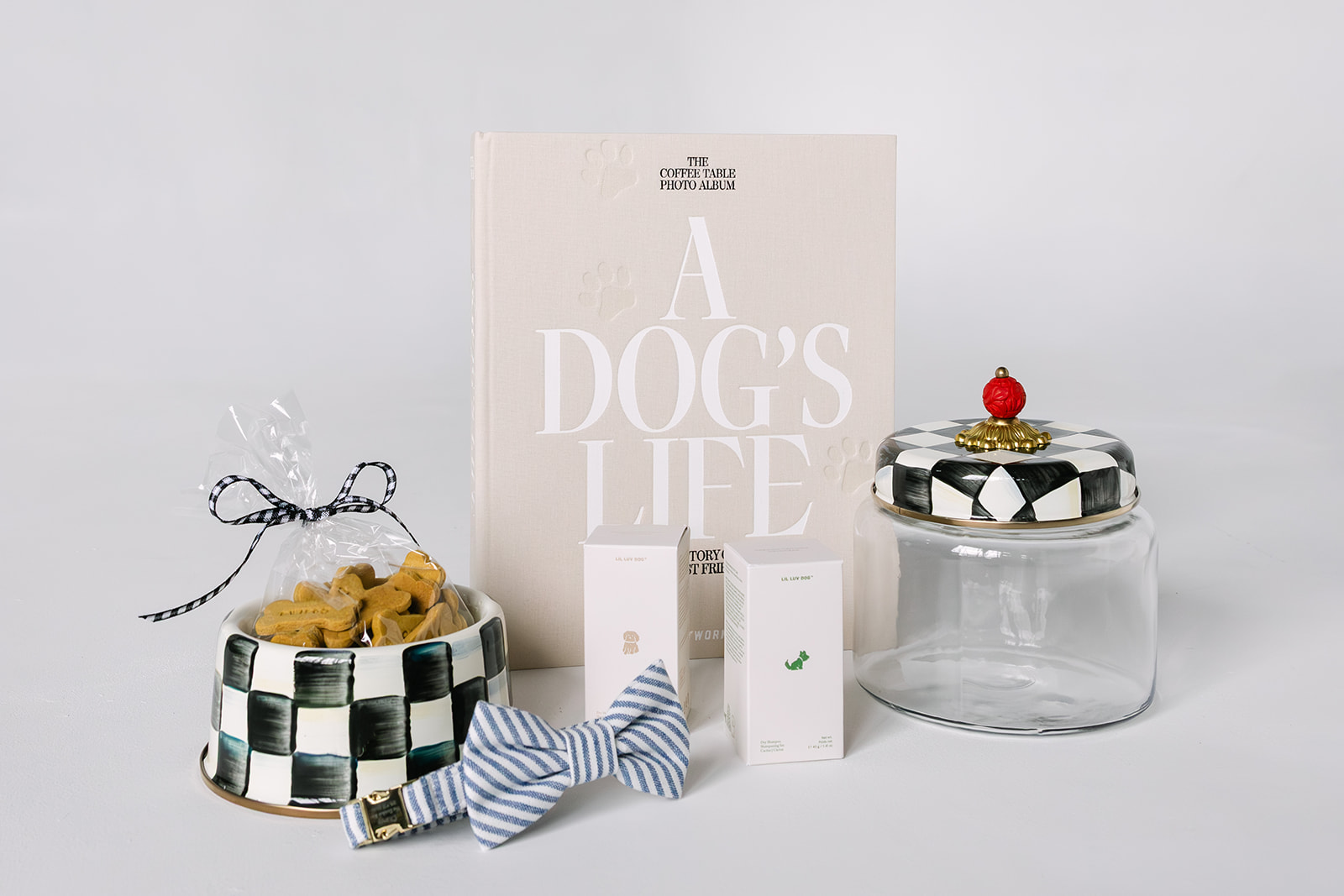 Collection of dog-themed items including a book titled 'A Dog's Life,' a black and white checkered dog bowl with packaged dog biscuits, two small product boxes, a blue and white striped bow tie collar, and an empty glass jar with a checkered lid topped with a red decorative knob.