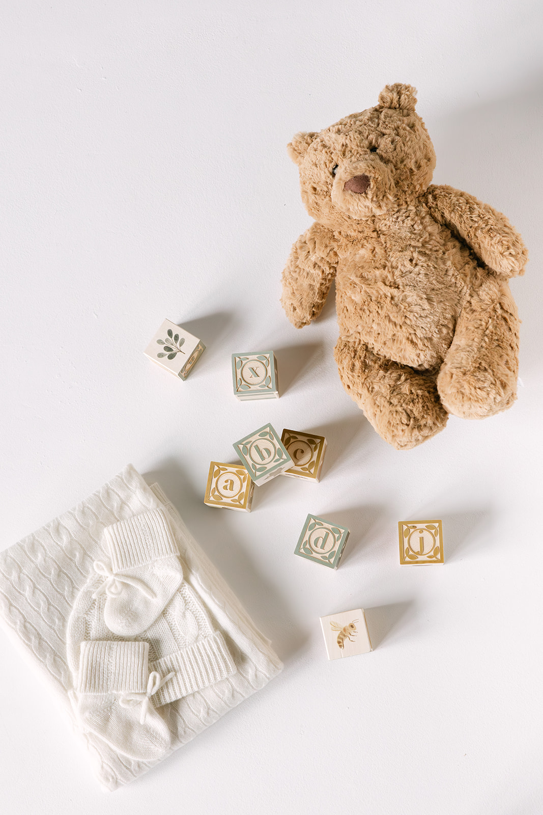 Light brown teddy bear, wooden alphabet blocks, and white knitted baby clothes including mittens and a hat on a white surface.