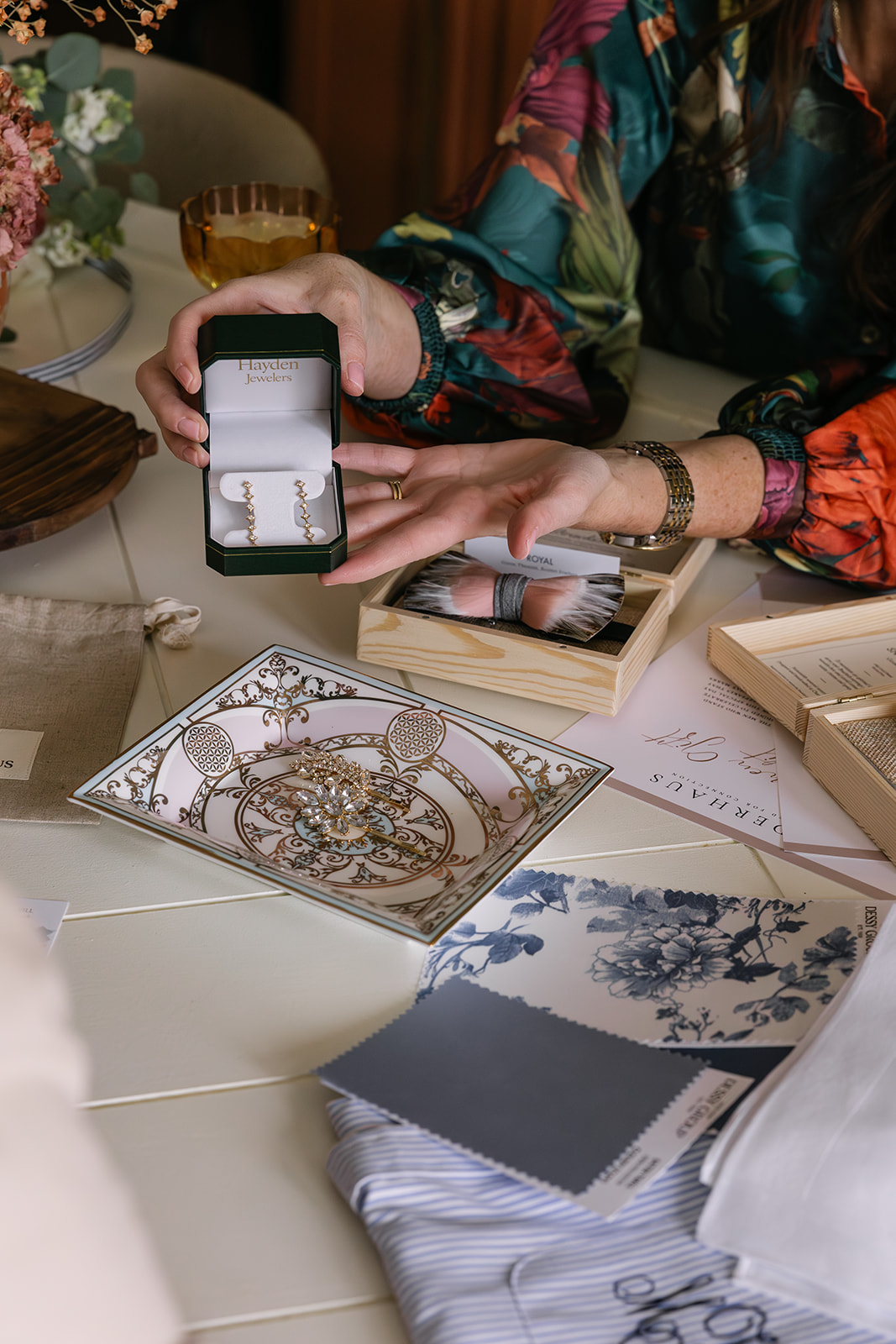 Person holding open green jewelry box showing gold and diamond earrings, surrounded by decorative plate with brooch, fabric swatches, and wooden boxes on a table.