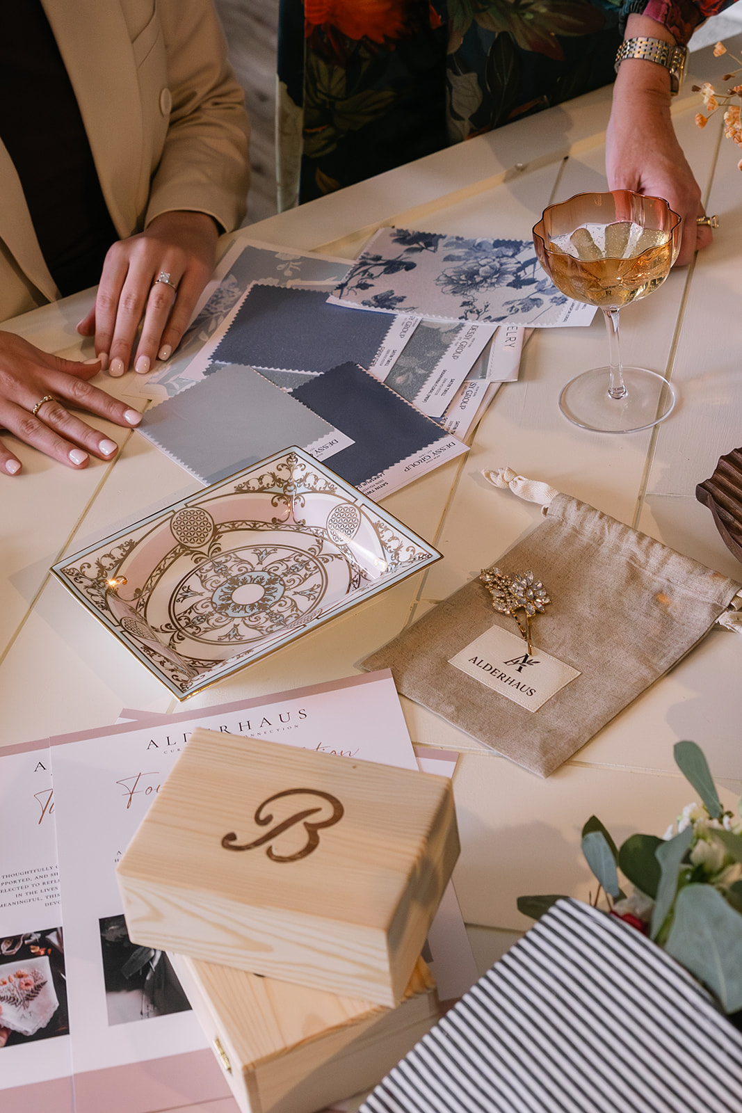 Two people at a table with fabric swatches, decorative plates, wood boxes, an Alderhaus jewelry pouch with a brooch, and a glass of beverage.